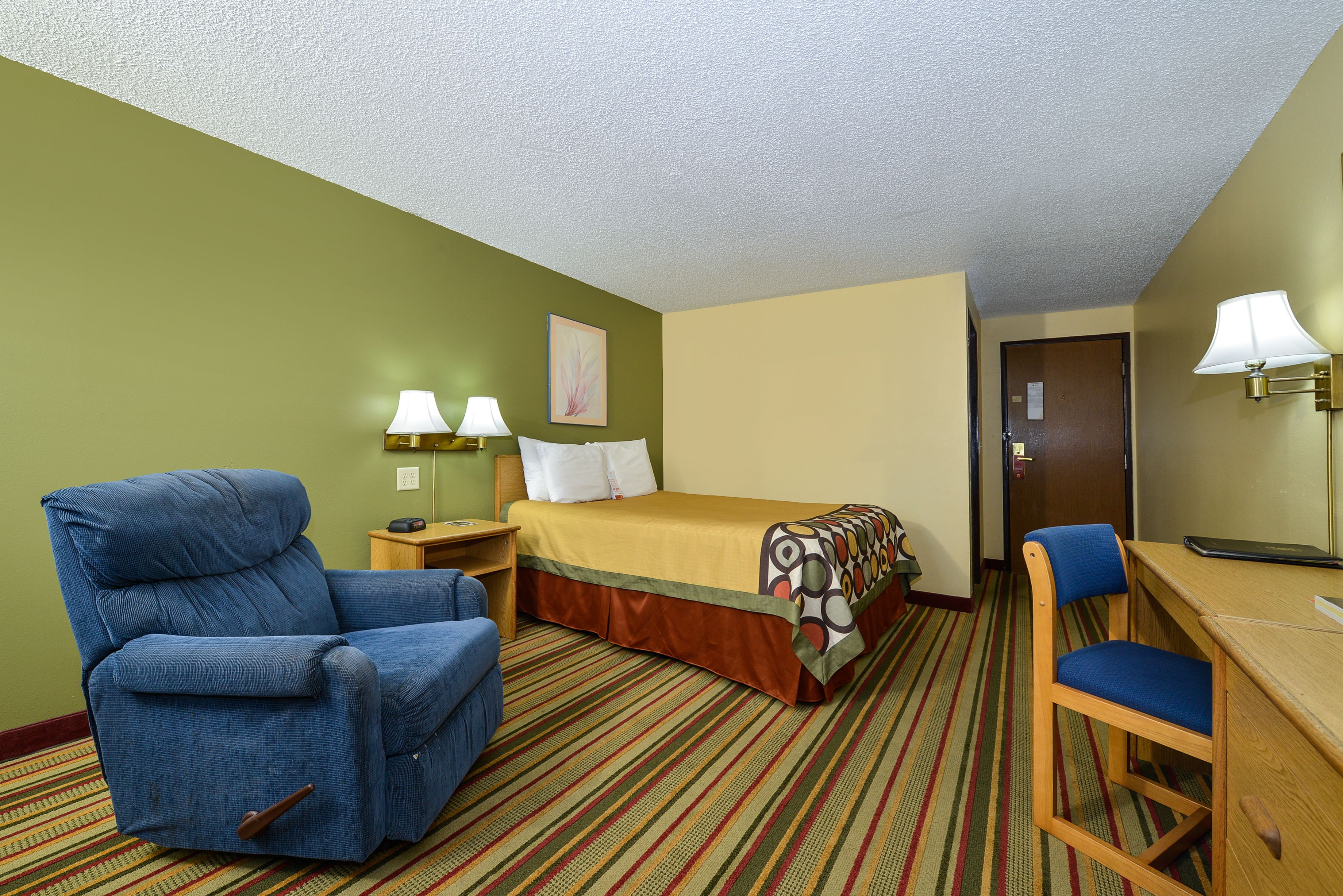 A hotel room with a bed, a blue armchair, a wooden desk, and a chair.