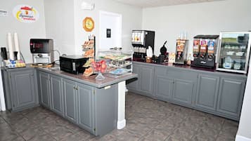 Un área de desayuno con una cafetera, una vitrina con pasteles y un dispensador de bebidas.