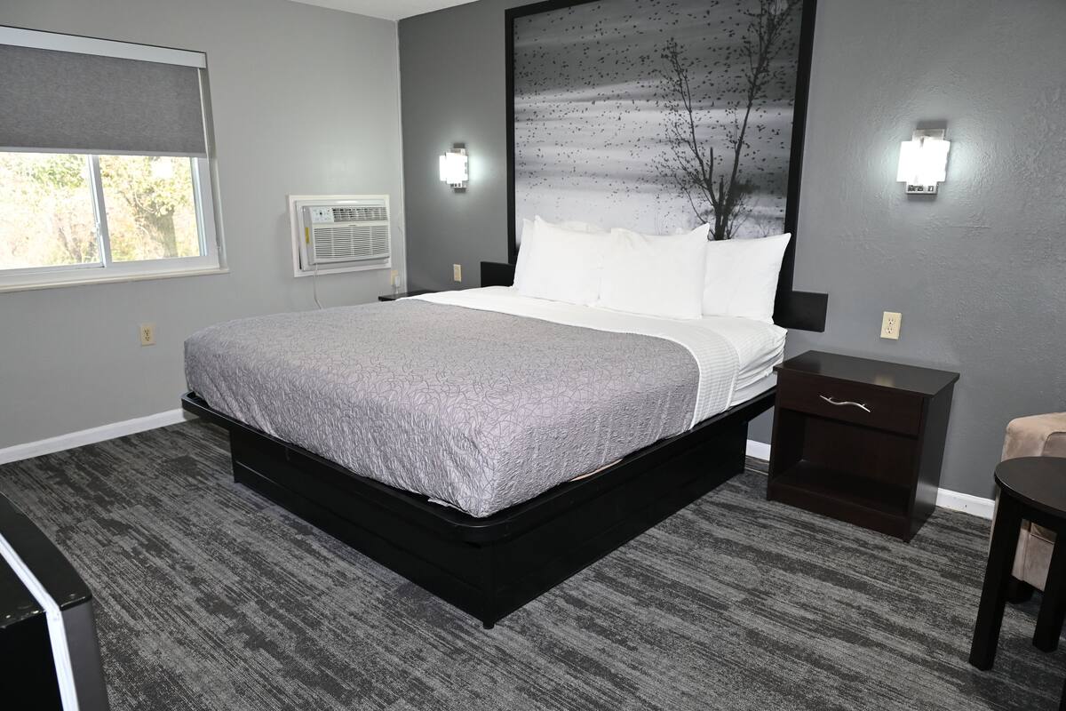 A modern bedroom with a large bed, a nightstand, a dresser, and a window with a view of trees.