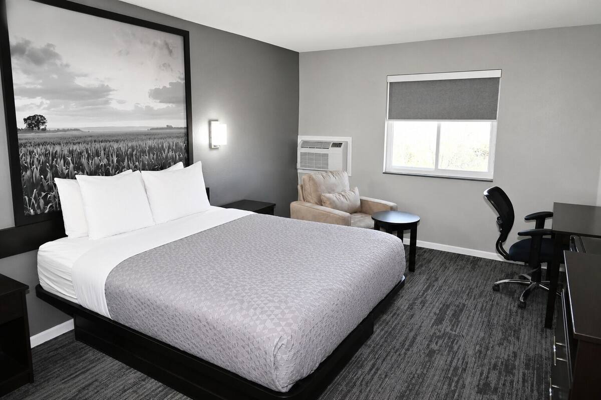 A hotel room with a large bed, a desk, a chair, a small table, a window with a view, and a wall-mounted air conditioning unit.