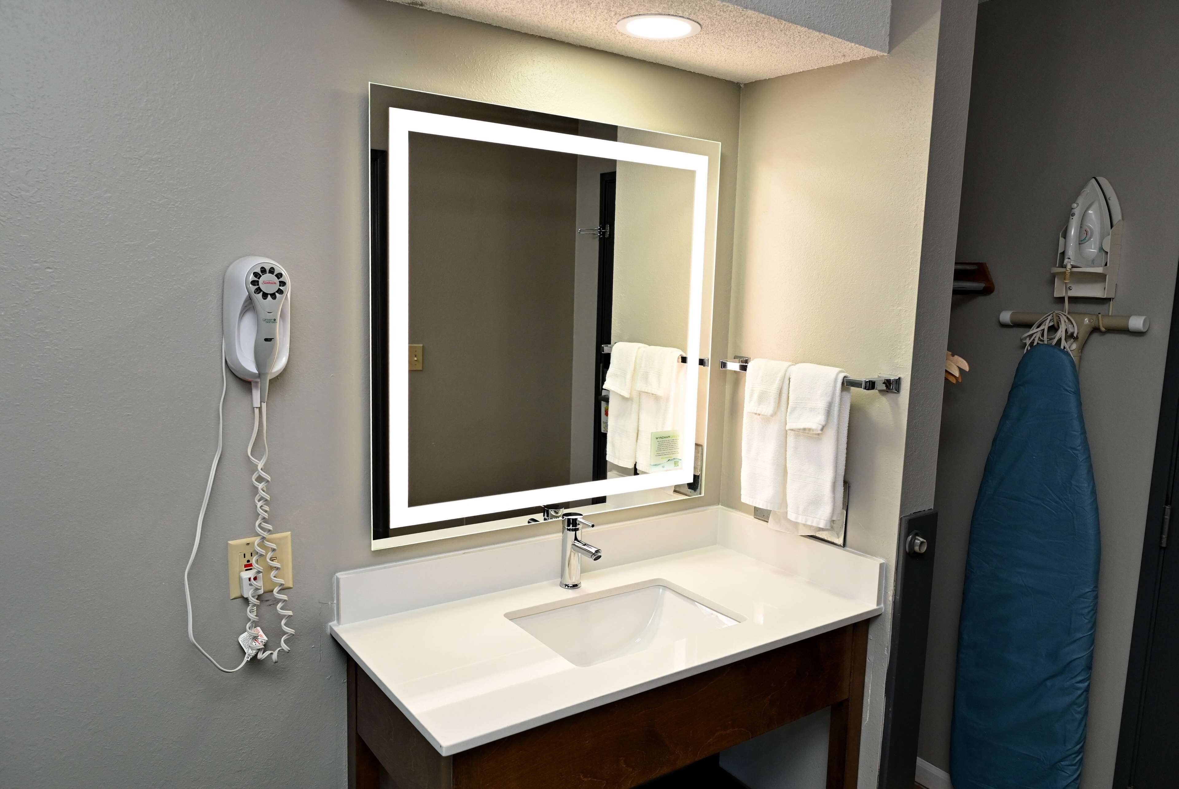 A bathroom with a white sink, a mirror with a light, a hairdryer, and a towel rack.