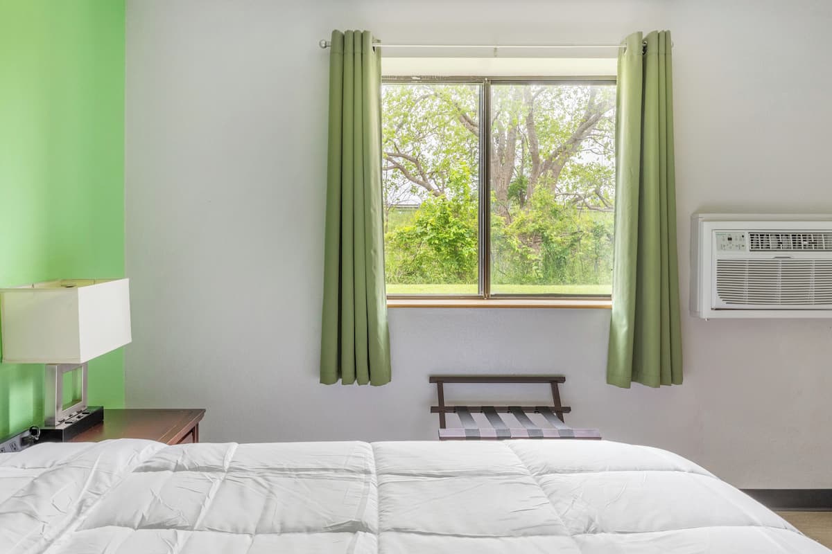 A room with a bed, a bench, a window with curtains, a wall-mounted air conditioner, and a lamp on a nightstand.