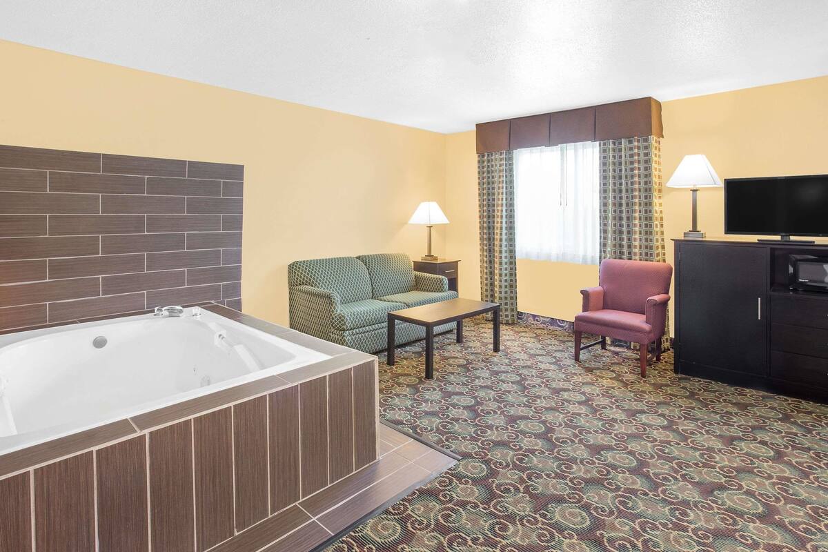 Une chambre d’hôtel avec une grande baignoire, un canapé à motifs verts, un fauteuil rose, une petite table et un téléviseur posé sur un meuble.