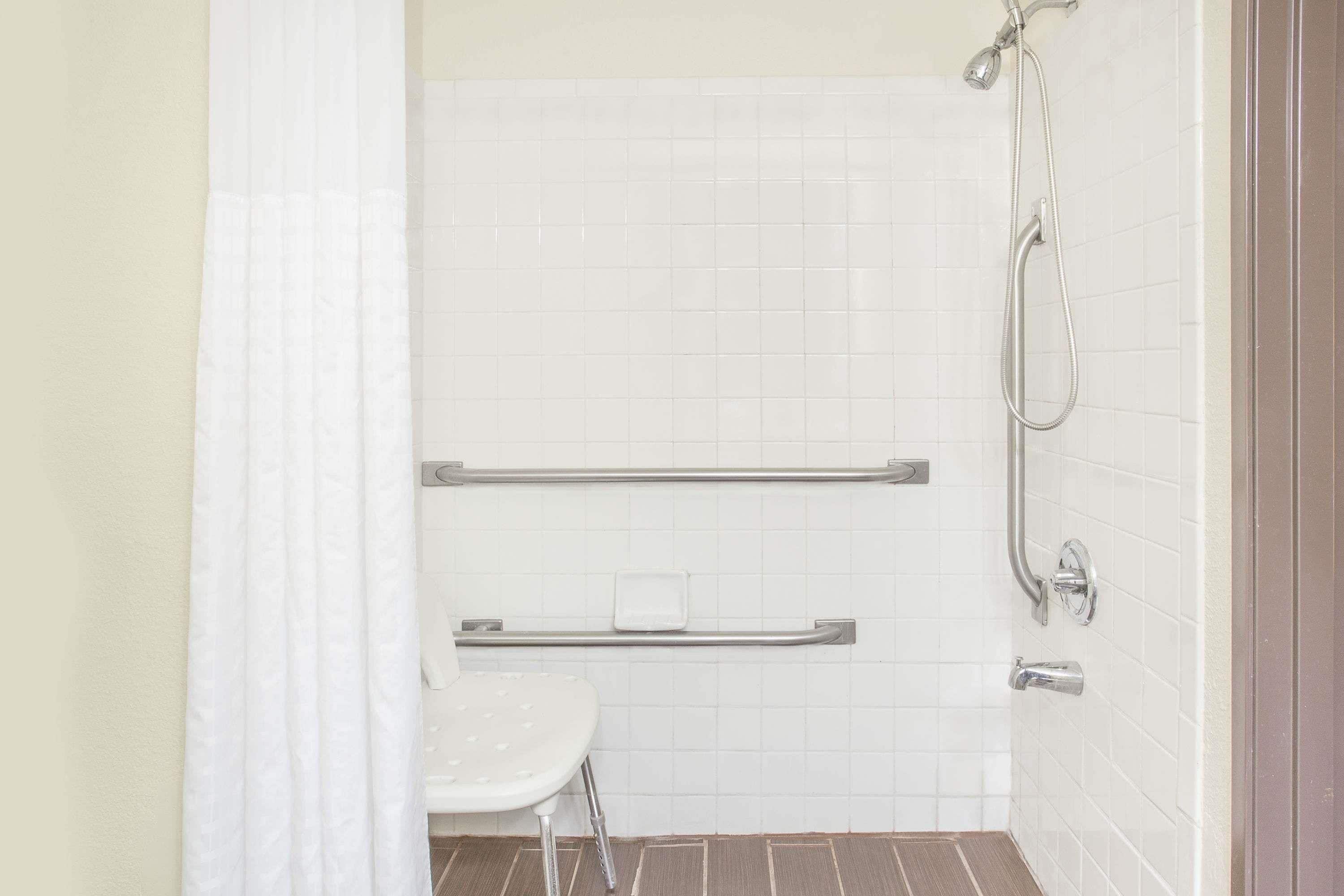 A bathroom with white tiled walls, a shower with a handheld showerhead, and a grab bar.