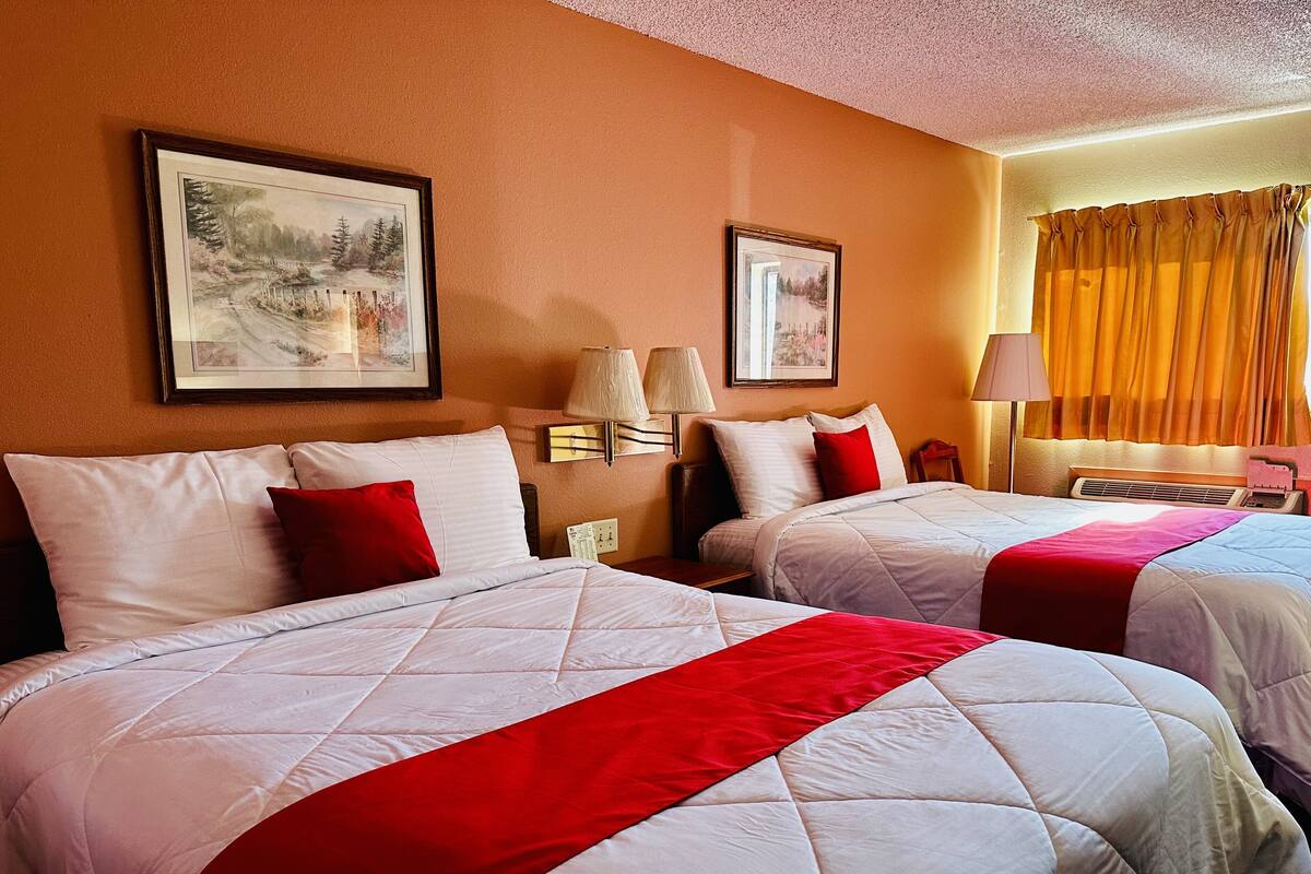 Habitación de hotel con dos camas, cada una con ropa de cama blanca y detalles en rojo, mesitas de noche con lámparas y cuadros en las paredes.