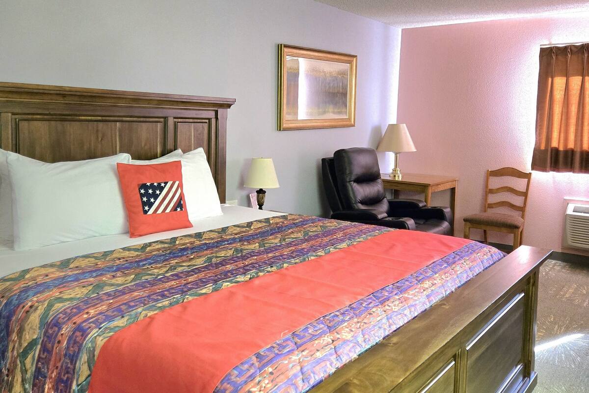 Habitación de hotel con una cama grande, cabecero de madera, escritorio con silla, lámpara y ventana con cortinas.