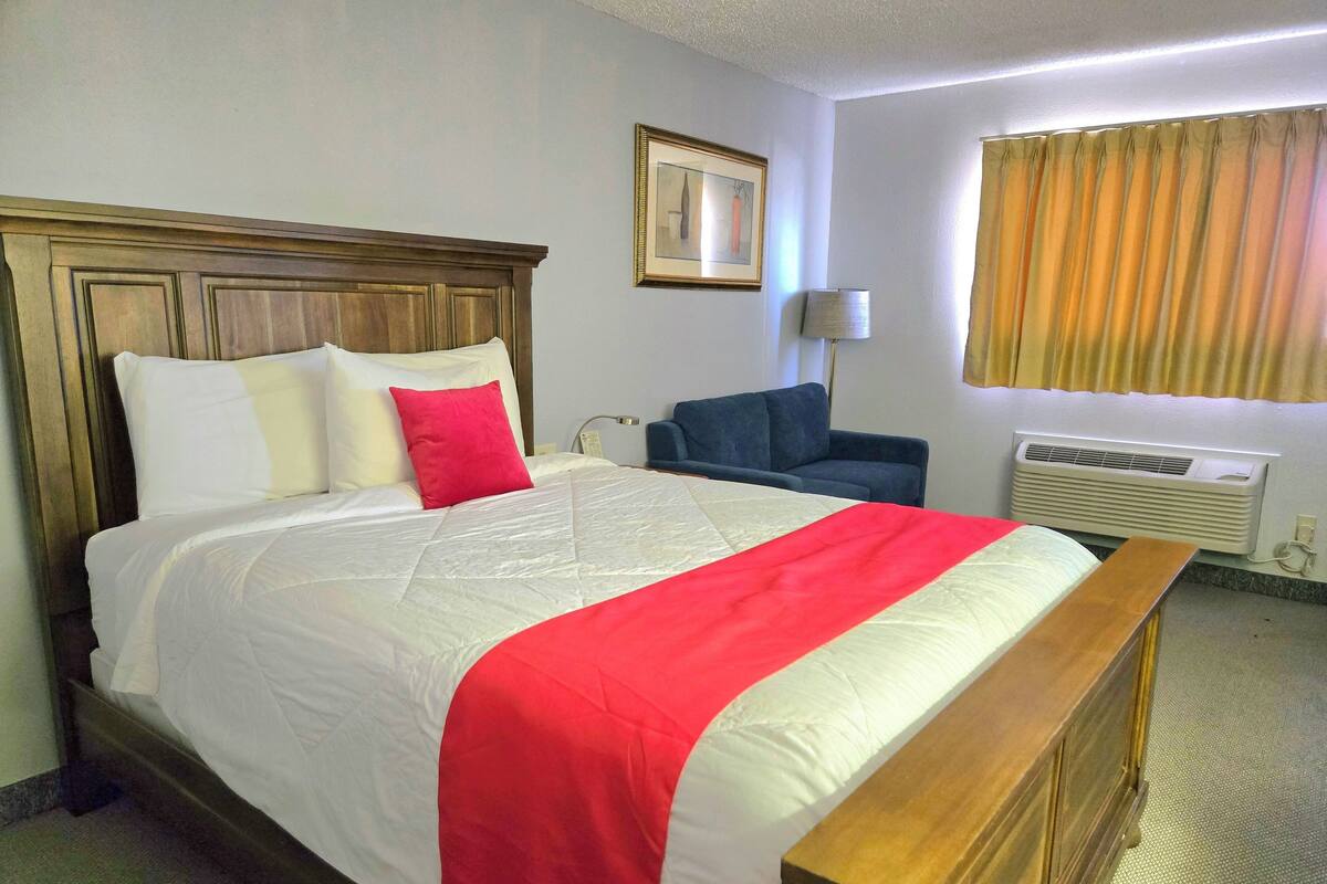 Habitación de hotel con cabecera de madera, una cama grande con ropa blanca y roja, un sillón azul, una lámpara de pie y una ventana con cortinas.