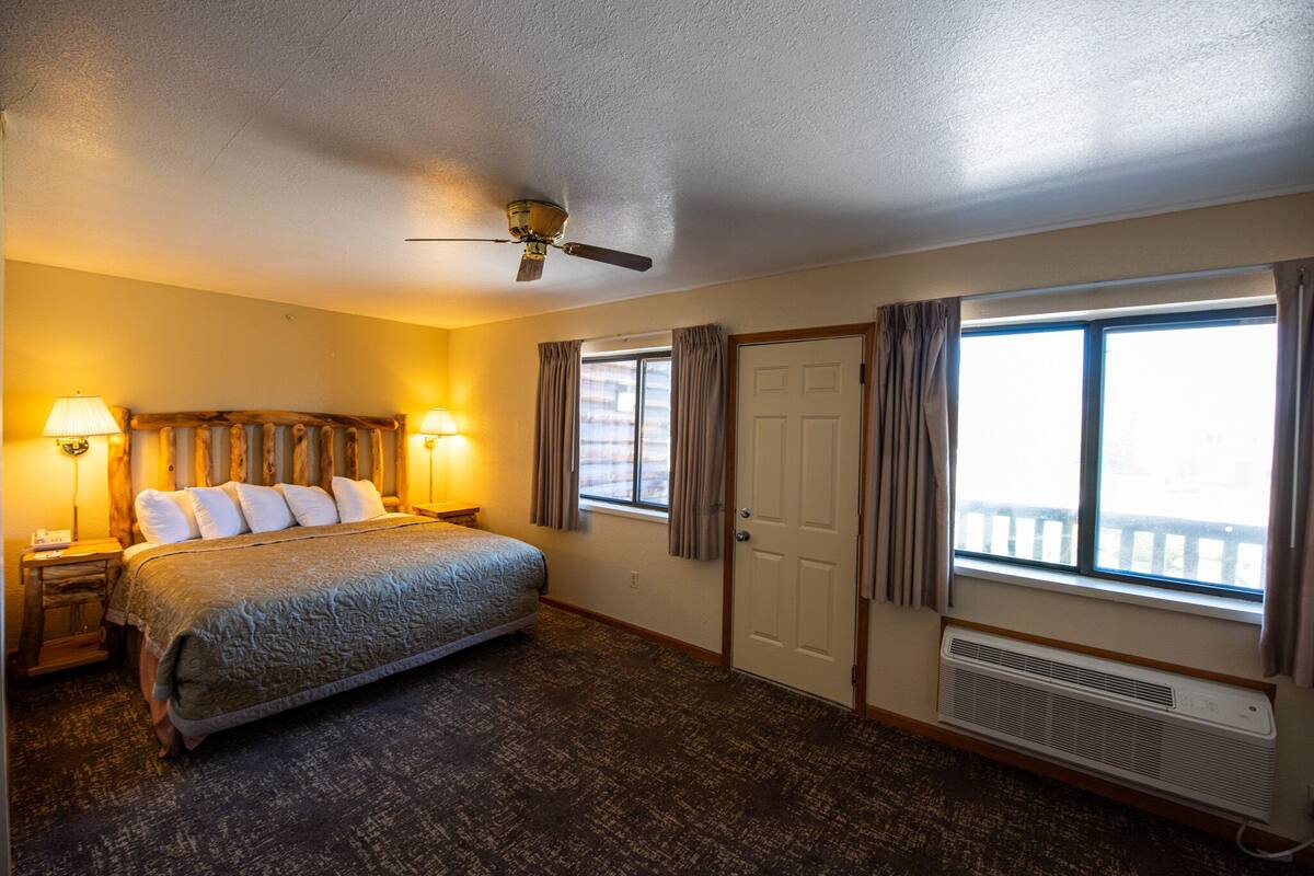 Une chambre d’hôtel avec un lit, deux lampes de chevet, un ventilateur de plafond, une fenêtre avec des rideaux et un radiateur.