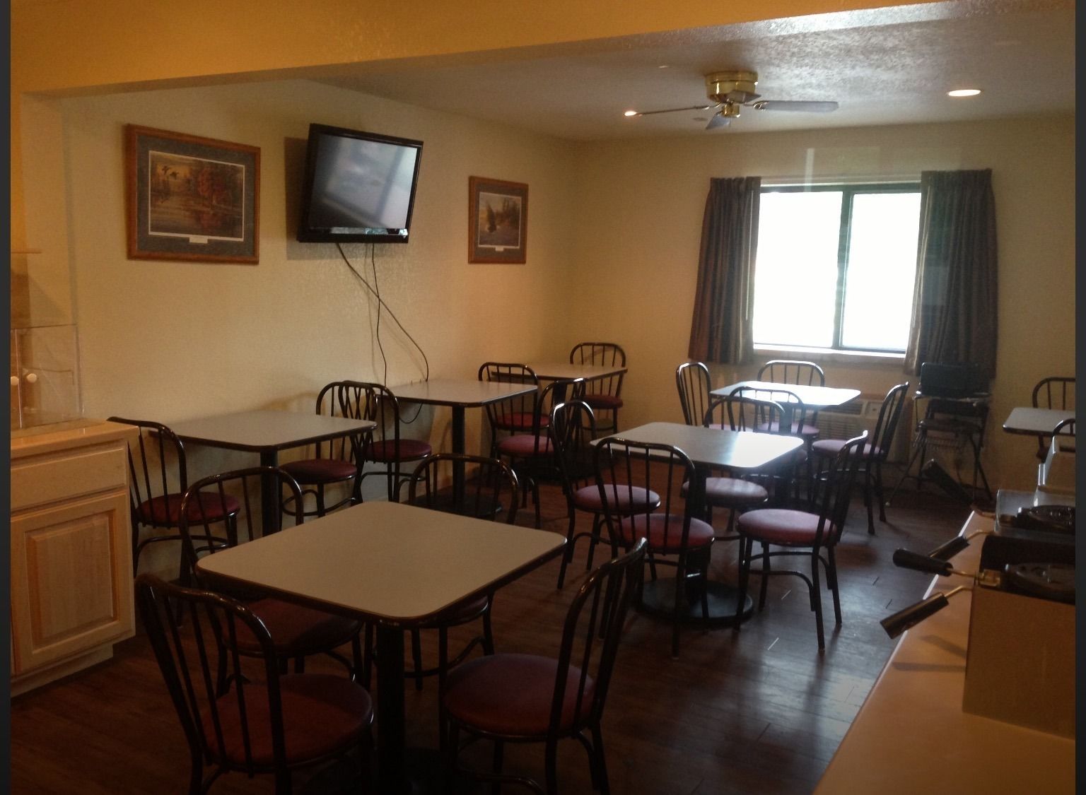 A dining area with multiple tables and chairs, a television mounted on the wall, and a window with curtains.