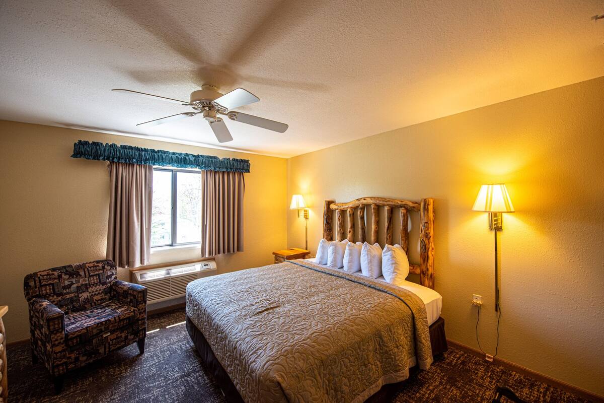 Une chambre d’hôtel avec un lit, une chaise, un ventilateur de plafond, une fenêtre avec des rideaux et deux lampes de chevet.