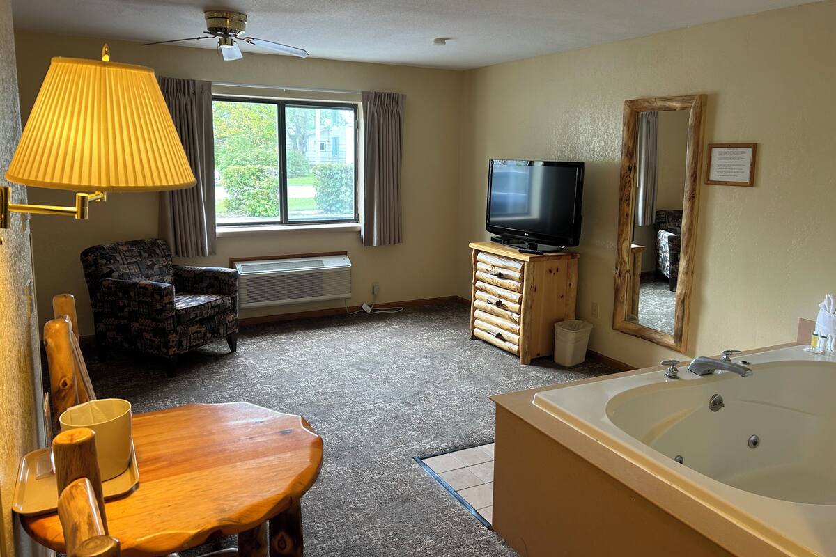 Une chambre d’hôtel avec une grande fenêtre, un ventilateur de plafond, un téléviseur posé sur un meuble en bois, une chaise, une table en bois avec deux tasses et une baignoire.