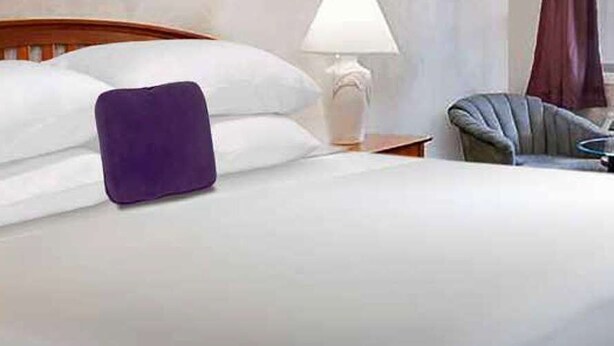 Un lit bien fait, avec du linge de lit blanc et un coussin violet, une tête de lit en bois, une lampe de chevet et une chaise avec une petite table ronde placée à côté du lit.