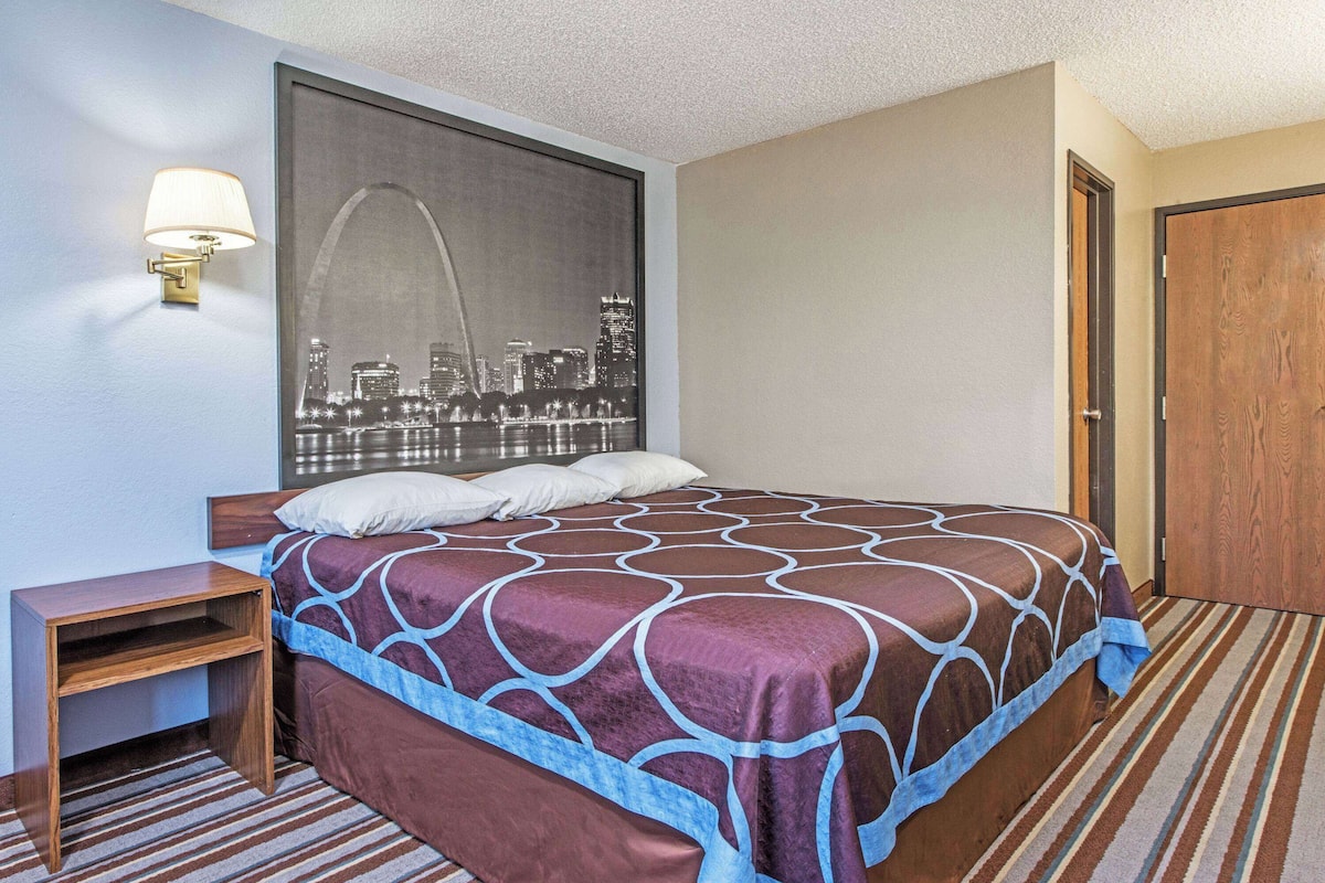 A hotel room with a large bed, a nightstand, a wall-mounted lamp, and a cityscape view.