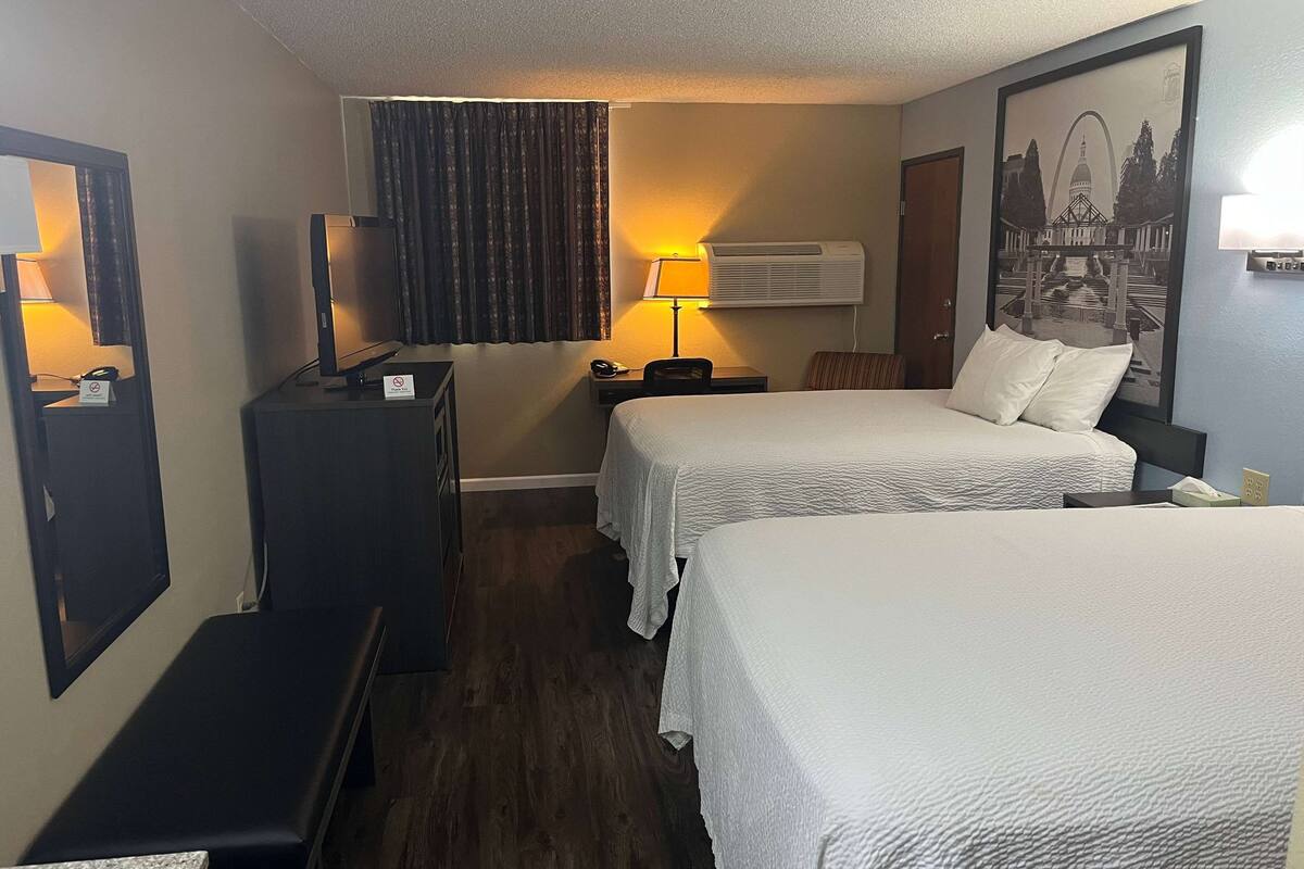 A hotel room with two beds, a desk, a chair, a lamp, an air conditioner, and a framed picture on the wall.