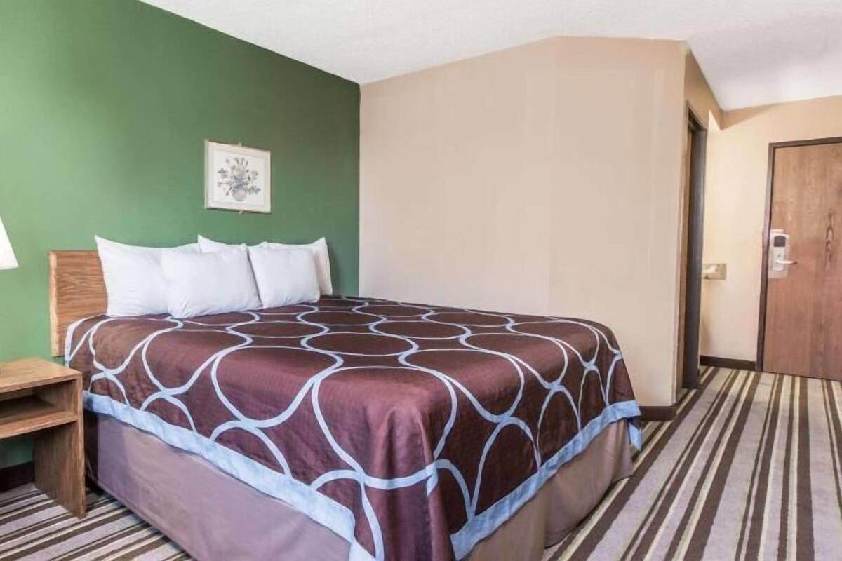 A hotel room with a bed, a nightstand, a lamp, a framed picture on the wall, and a door.