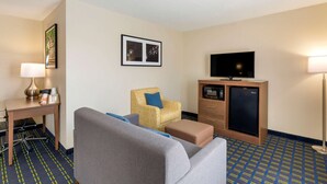 A hotel room with a sofa, armchair, desk with lamp, television, mini-fridge, and patterned carpet. - Quality Inn Lee - Lenox South (Lee)