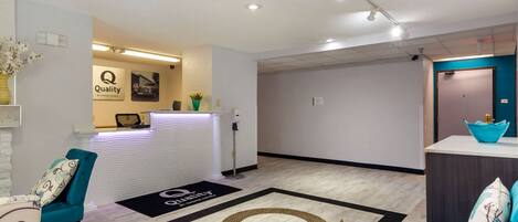 A hotel reception area with a reception desk, a teal chair, a sofa, and a large logo rug.