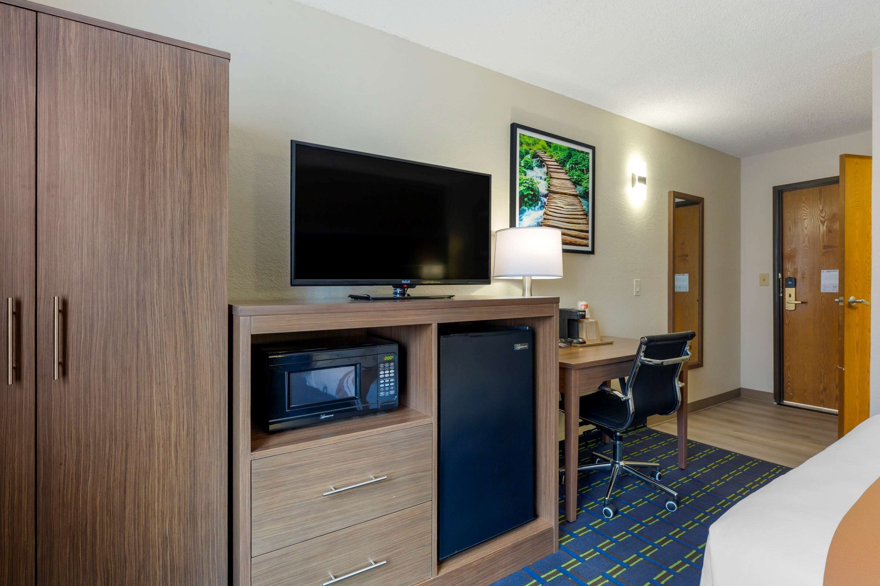 A hotel room with a wooden wardrobe, a flat-screen TV, a microwave, a refrigerator, a desk with a chair, and a bed.