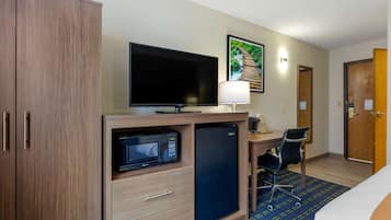A hotel room with a wooden wardrobe, a flat-screen TV, a microwave, a refrigerator, a desk with a chair, and a bed.