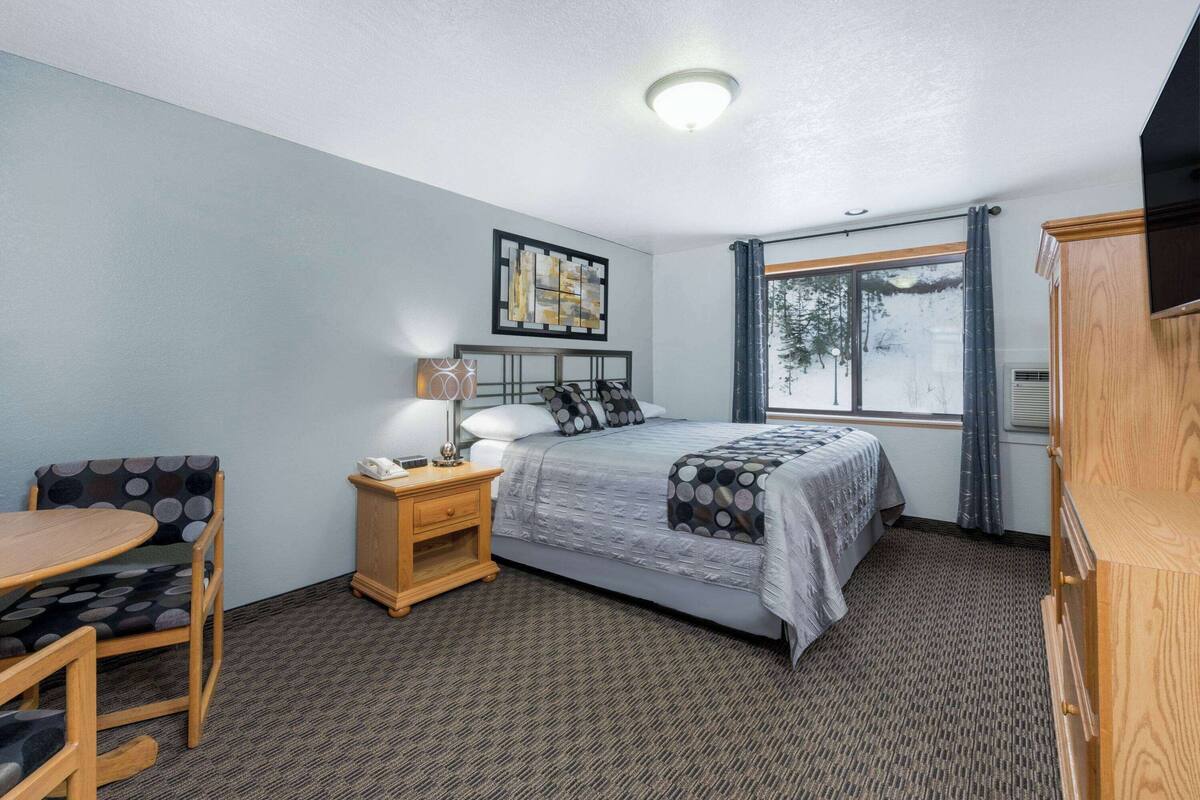 Un dormitorio con cama, mesita de noche, silla, mesa, ventana con cortinas y un cuadro en la pared.