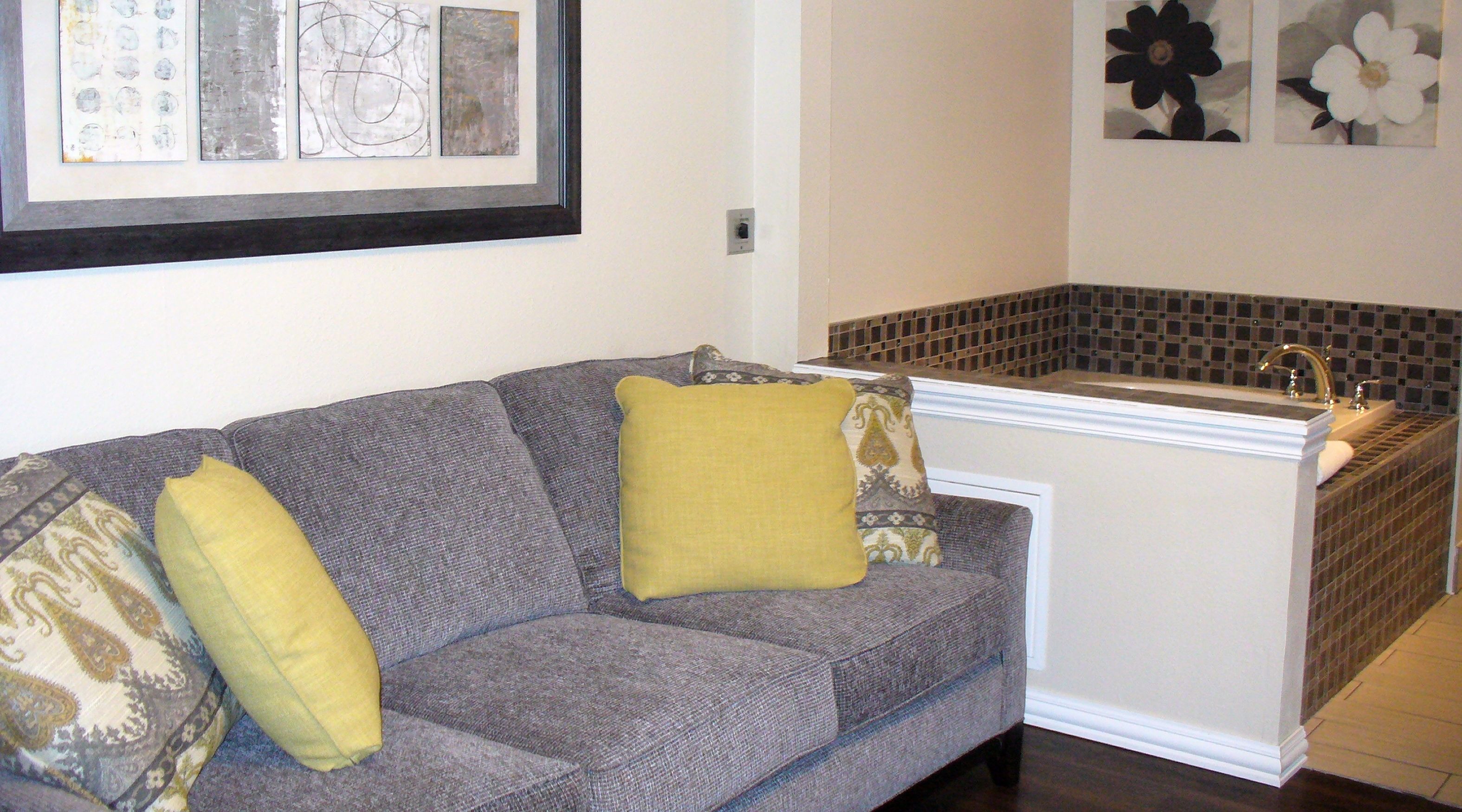 A grey sofa with yellow and patterned cushions, a tiled shower area, and framed artwork on the wall.