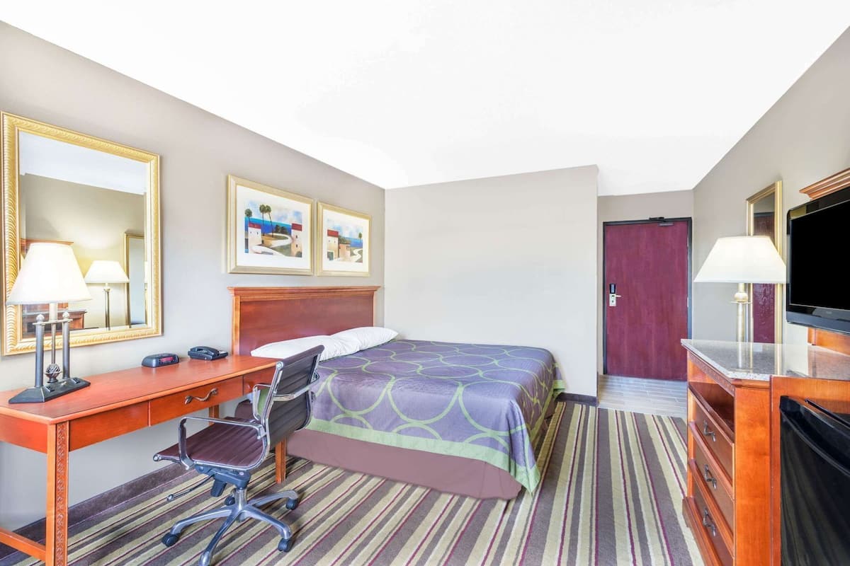 Une chambre d’hôtel avec un lit, un bureau, une chaise, deux lampes, une télévision et une porte.