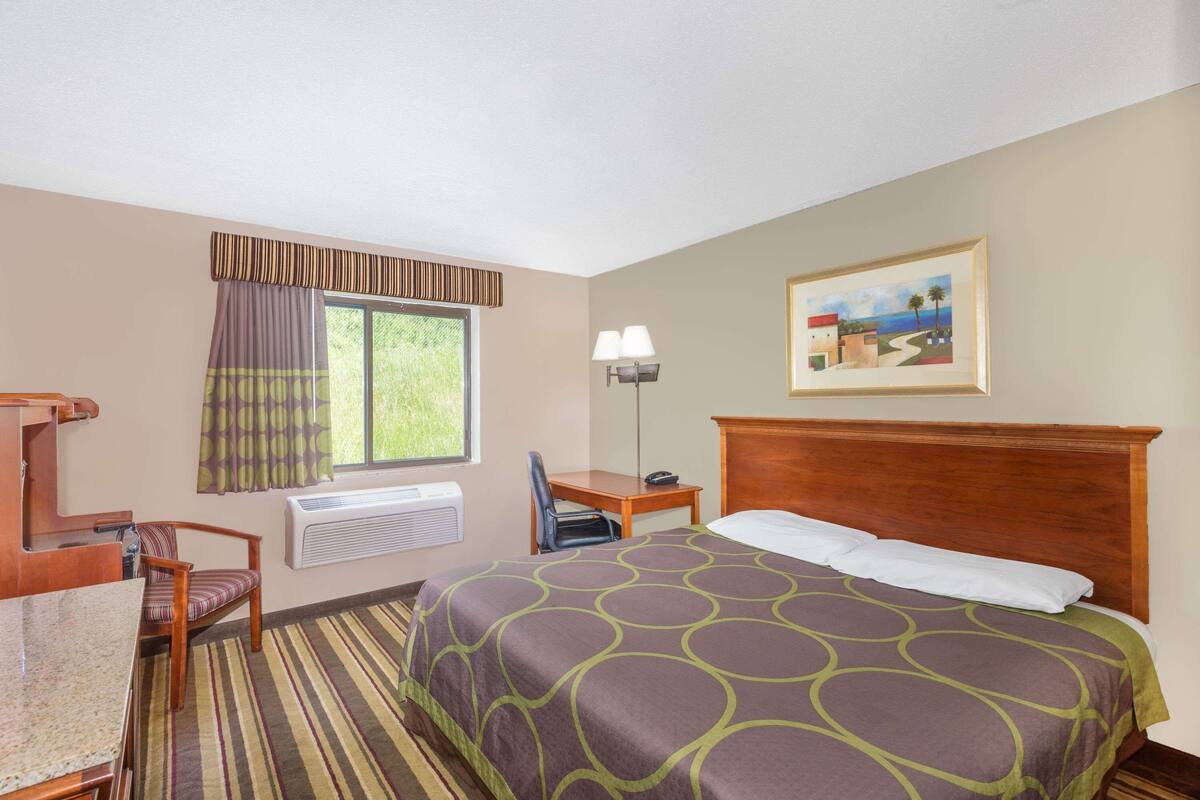 A hotel room with a bed, a desk, a chair, a window with curtains, a painting on the wall, and a radiator.