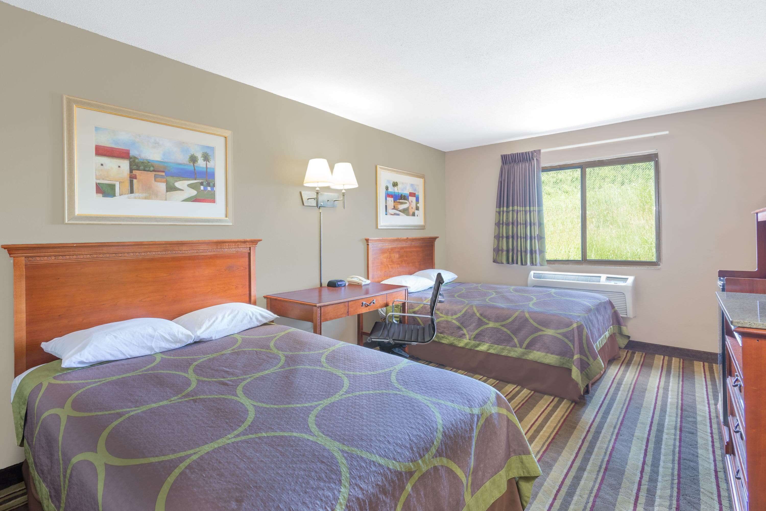 A hotel room with two beds, a desk, a chair, a lamp, and a window with curtains.