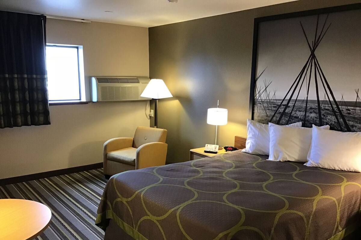 Una habitación de hotel con cama, sillón, mesita de noche, lámpara y ventana con cortinas.
