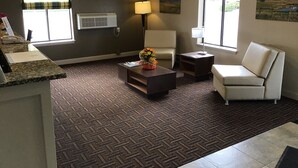 Habitación de hotel con sofá, sillón, mesa de centro y escritorio con teléfono. Hay dos ventanas con cortinas, un aire acondicionado instalado en la pared y un cuadro en la pared.