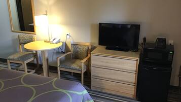 Una habitación de hotel con cama, escritorio con lámpara, dos sillas, televisor, microondas y cómoda.