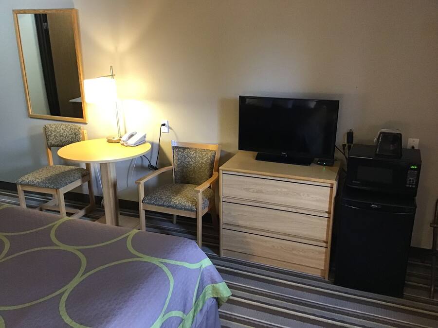 Una habitación de hotel con cama, escritorio con lámpara, dos sillas, televisor, microondas y cómoda.