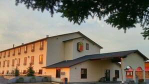 Un hotel con un edificio grande y una estructura más pequeña adosada, ambos con múltiples ventanas y un estacionamiento al frente.