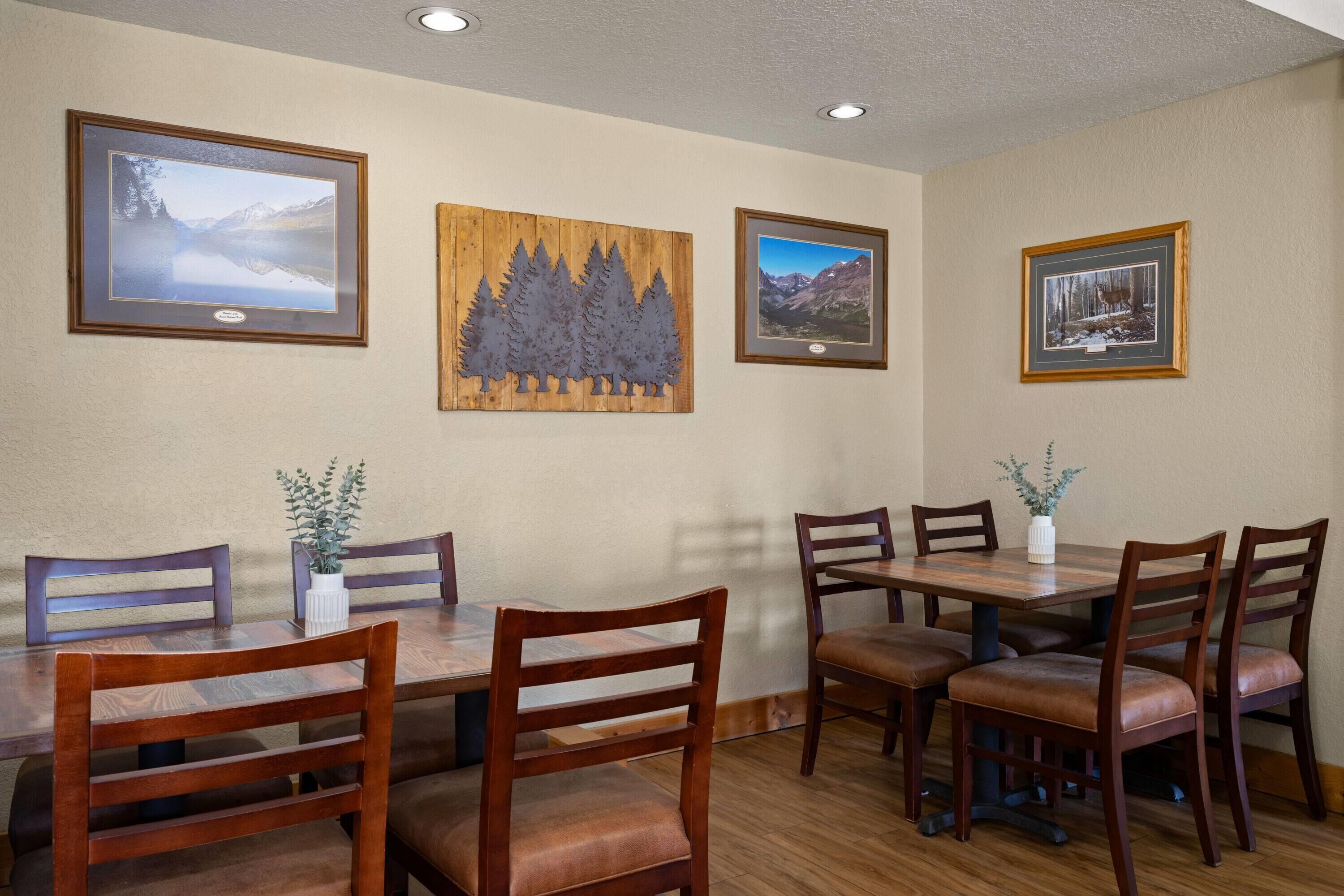 A dining area with wooden tables and chairs, framed pictures on the wall, and a potted plant on the table.