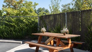 A wooden picnic table with two flower pots on top, set against a chain-link fence and surrounded by greenery. - Super 8 by Wyndham Kalispell Glacier National Park (Kalispell)