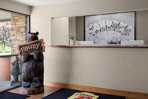 A reception desk with a framed welcome sign, a wooden bear sculpture holding a sign, and a small potted plant. - Super 8 by Wyndham Kalispell Glacier National Park (Kalispell)