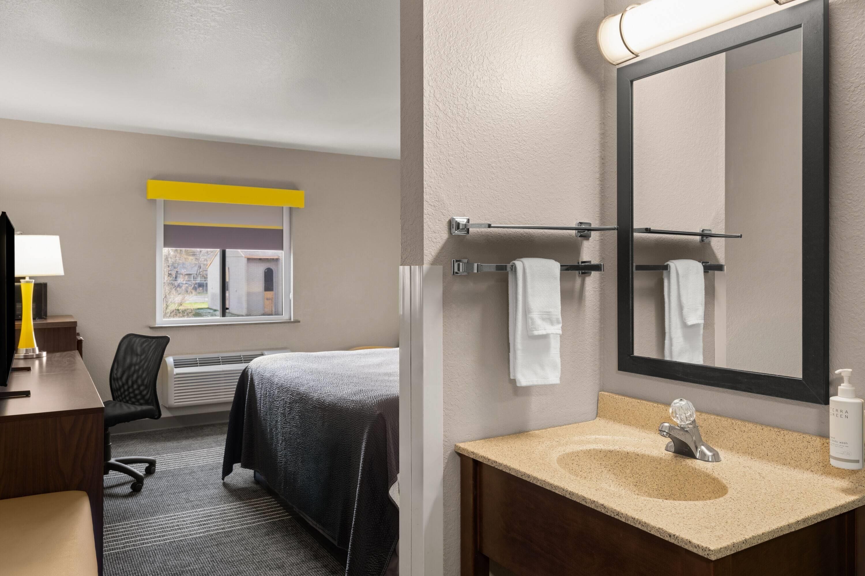 Habitación de hotel con cama, escritorio con lámpara, baño con lavamanos y espejo, y ventana con vistas.