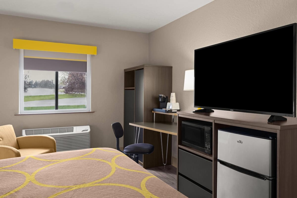 Quarto de hotel com uma cama, escrivaninha com computador, televisão, mini-frigorífico e janela com vista para um lago.
