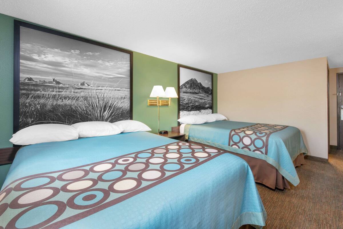 A hotel room with two beds, each with a blue bedspread featuring a circular pattern. There is a wall-mounted lamp between the beds, and the room has a green accent wall with a large black-and-white photograph.