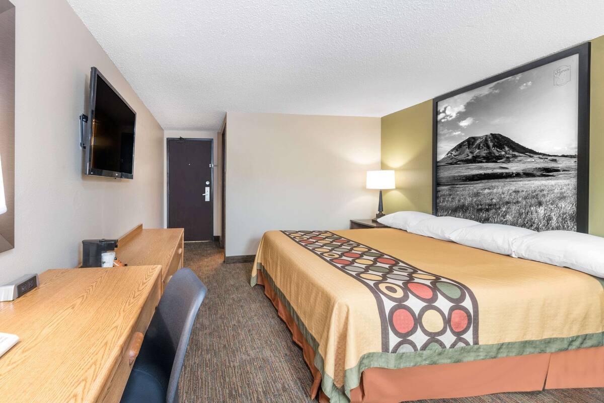 A hotel room with a bed, a desk, a chair, a TV mounted on the wall, and a large framed picture of a mountain landscape.