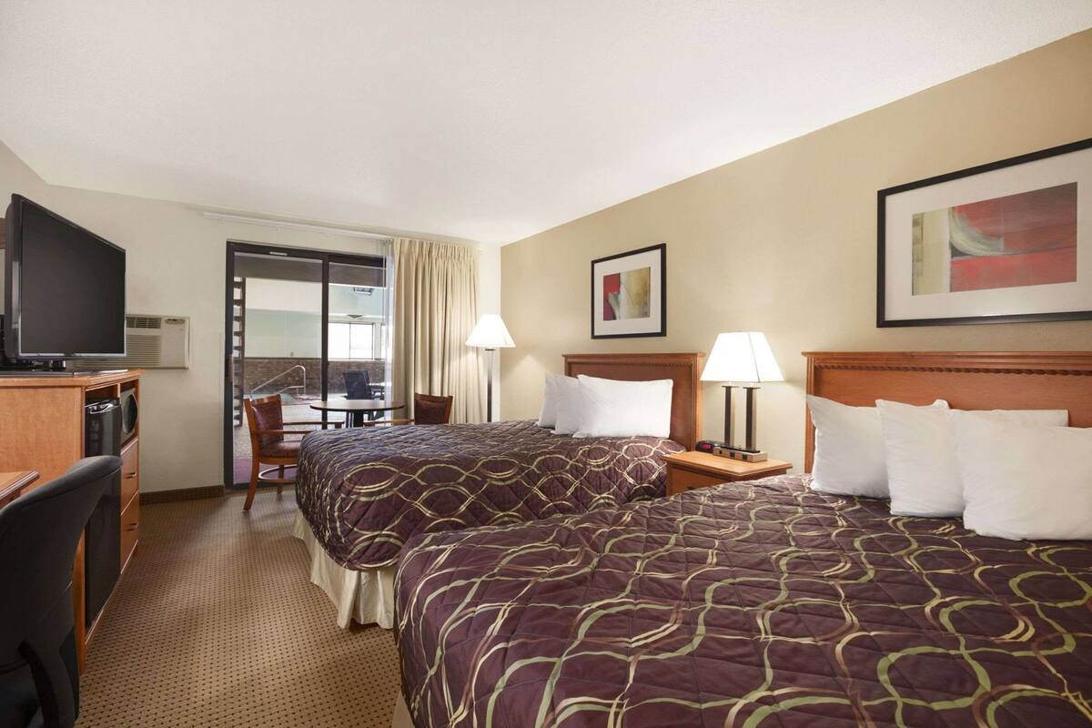 A hotel room with two beds, a desk with a television, a chair, a table, and a window with curtains.
