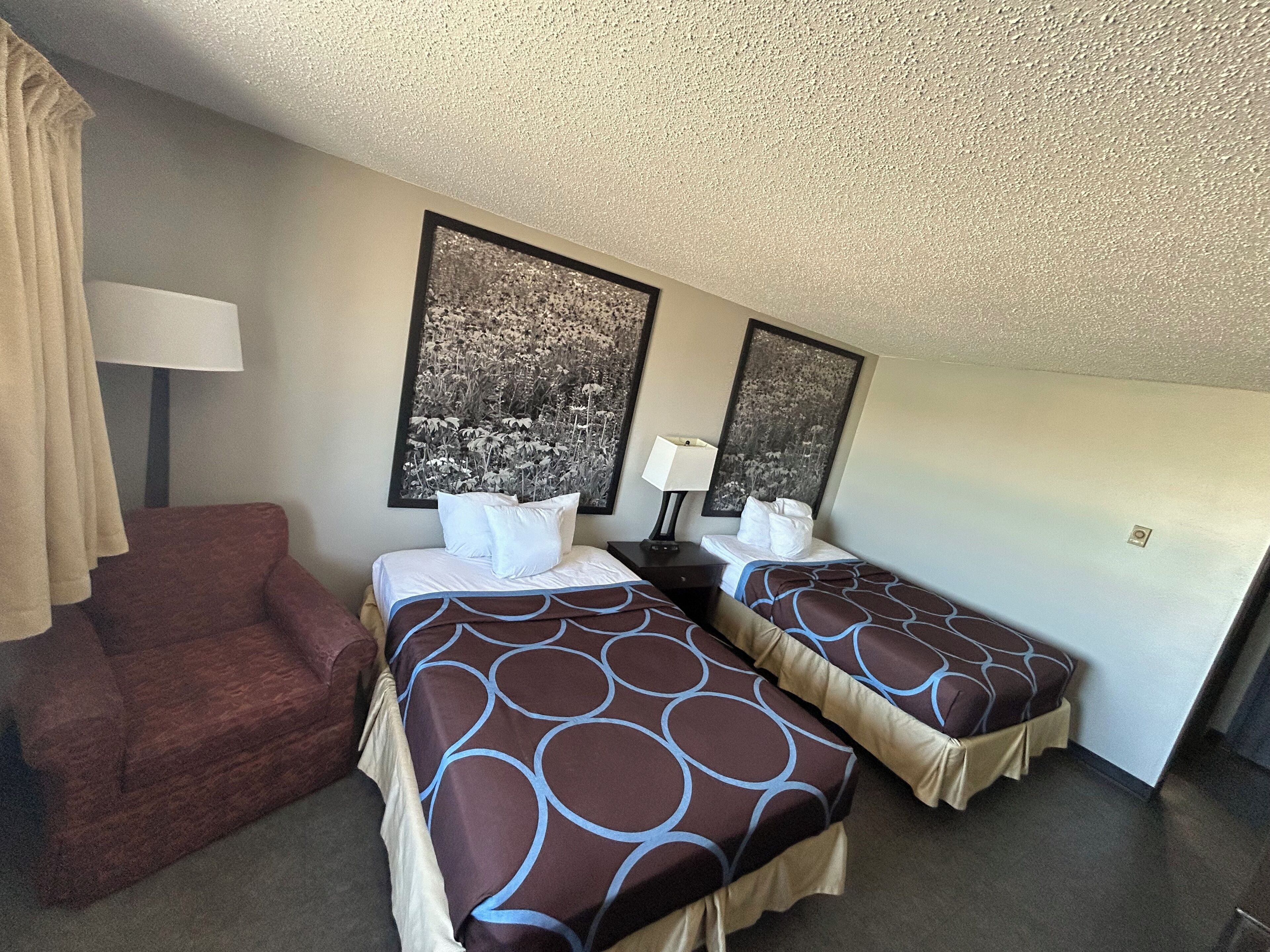 A hotel room with two beds, a red armchair, a lamp, and two framed pictures on the wall.
