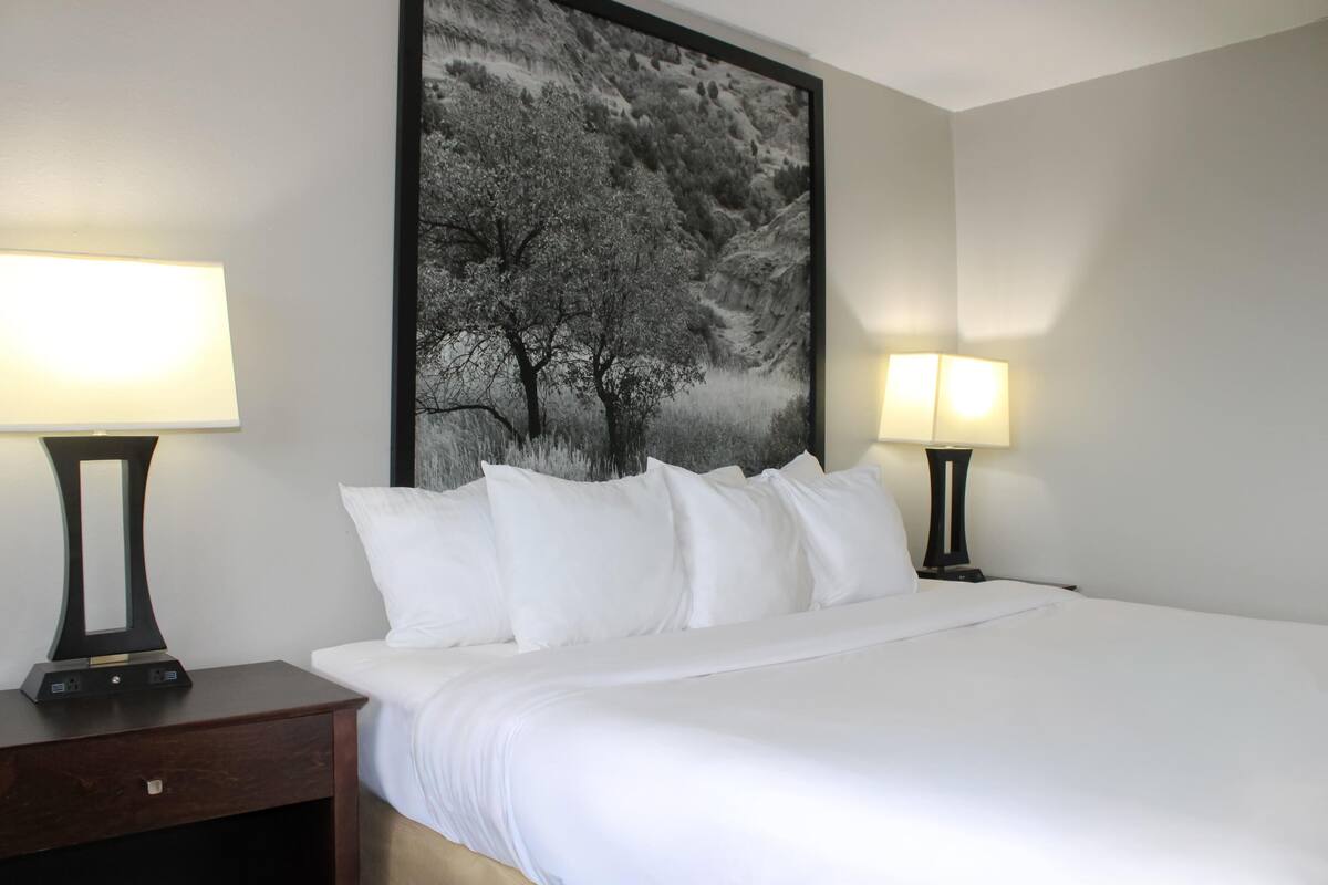 A neatly made bed with white linens, two bedside lamps, a dark wooden nightstand with a drawer, and a large black and white landscape painting on the wall.