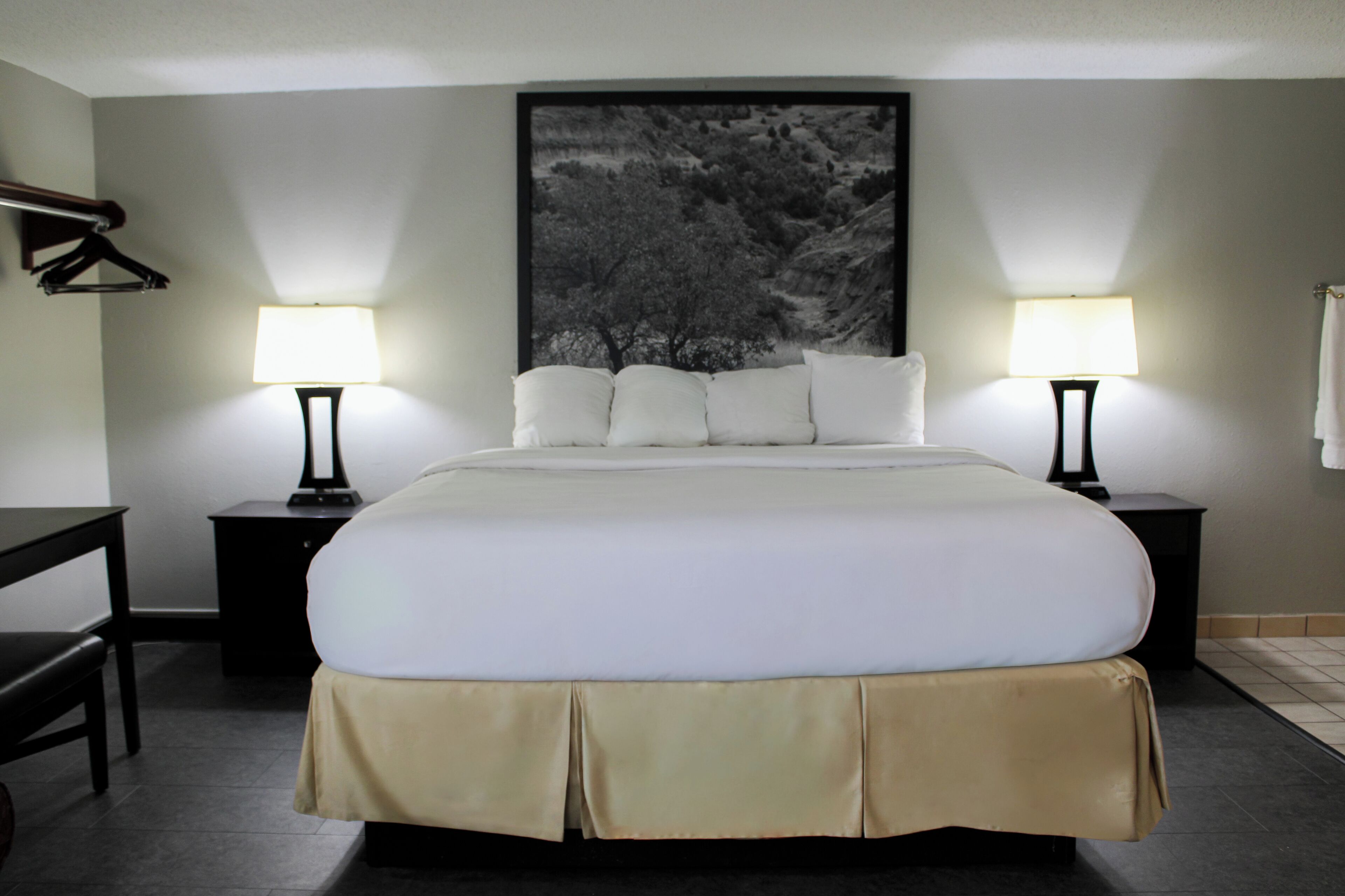 Una cama bien hecha con sábanas blancas y falda color beige, flanqueada por dos mesitas de noche con lámparas, una obra de arte con marco negro sobre la cama, y un escritorio de madera oscura con una silla a la izquierda.