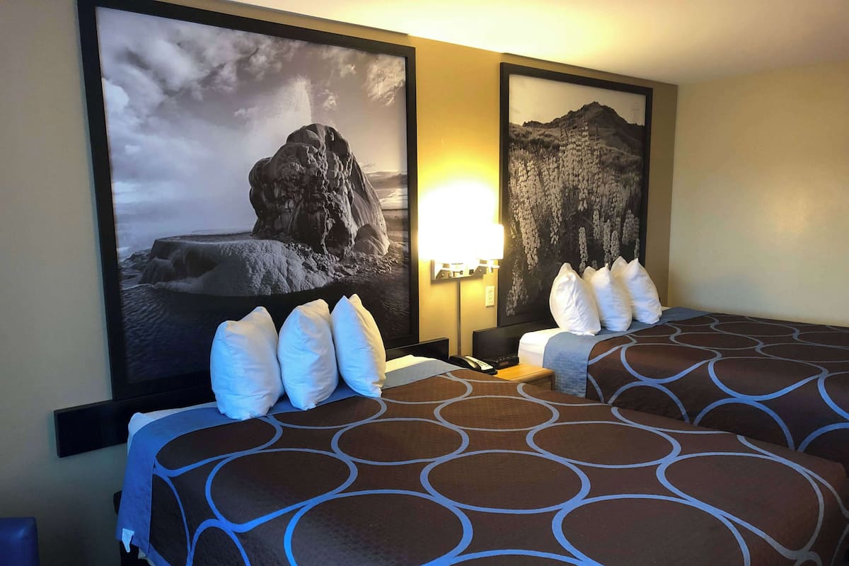 Ein Hotelzimmer mit zwei Betten, jeweils mit einem gemusterten Überwurf und weißen Kissen. An den Wänden hängen Bilder in Rahmen, es gibt eine Nachttischlampe und einen Flachbildfernseher.