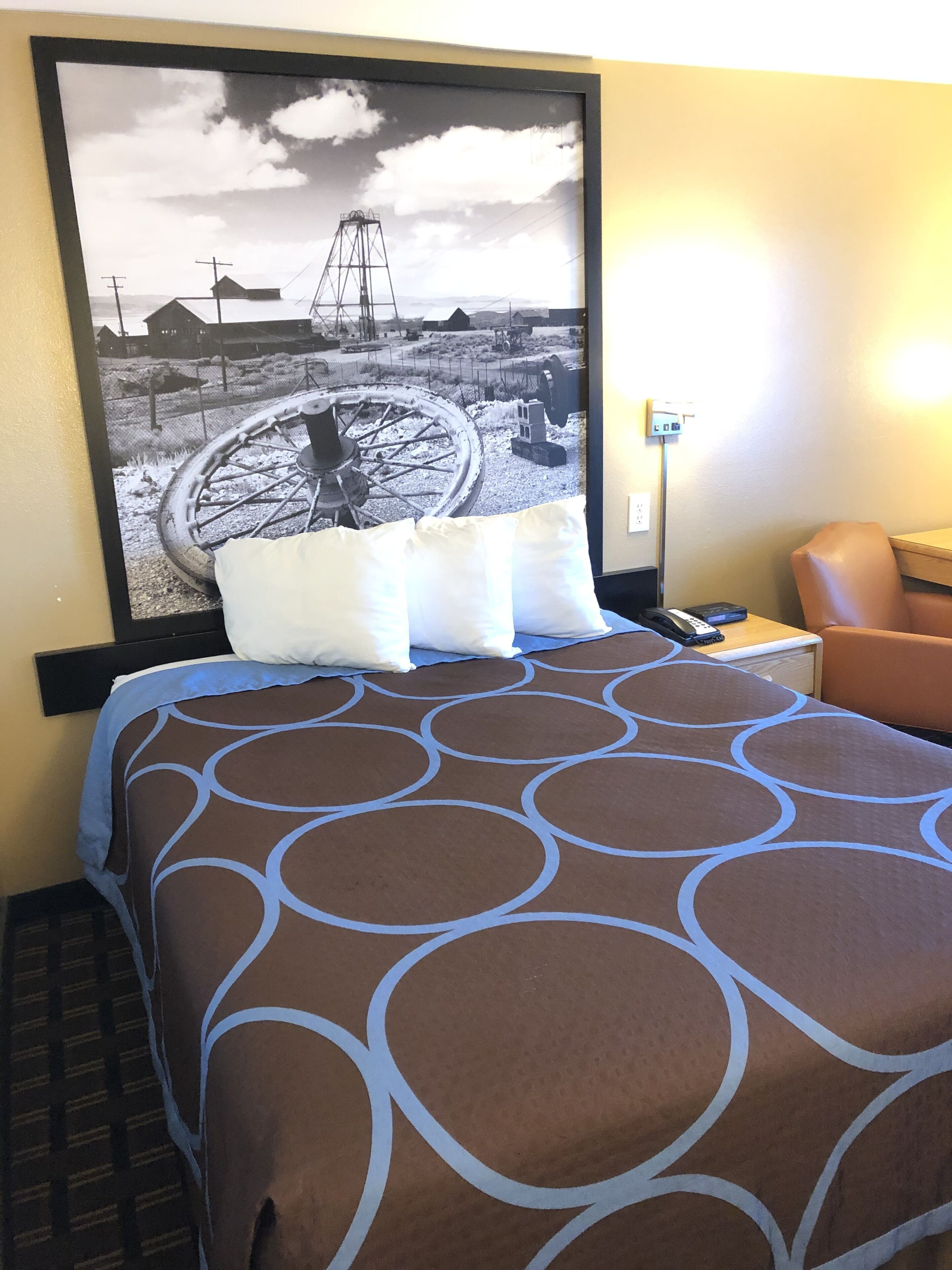 A hotel room with a large bed, a chair, a desk with a phone, and a wall-mounted picture of a mining scene.