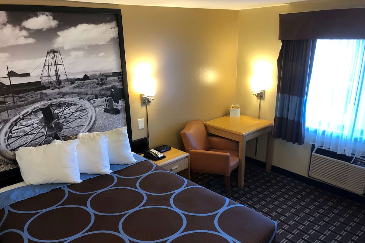 Ein Hotelzimmer mit Bett, Schreibtisch, Stuhl, Lampe und einem fenster mit Vorhängen.
