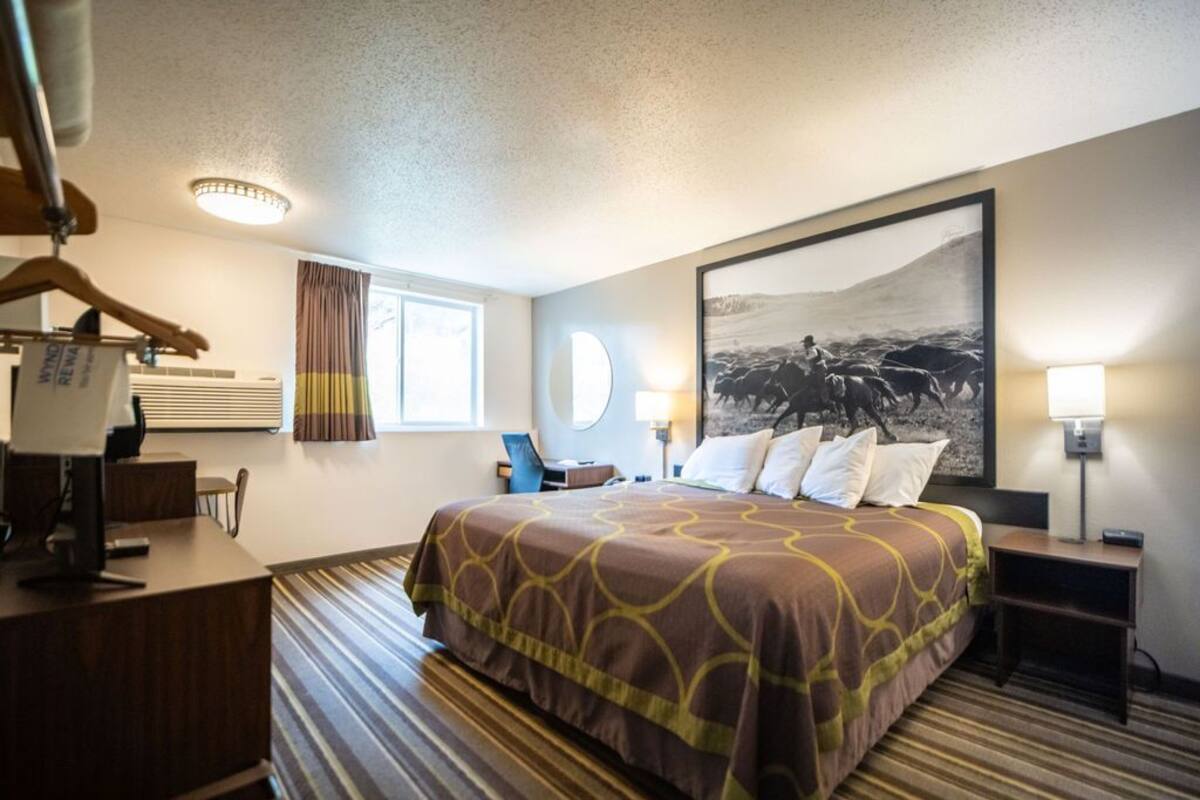 Een hotelkamer met een groot bed, een bureau met een computer, een stoel, een raam met gordijnen en een aan de muur hangend kunstwerk.