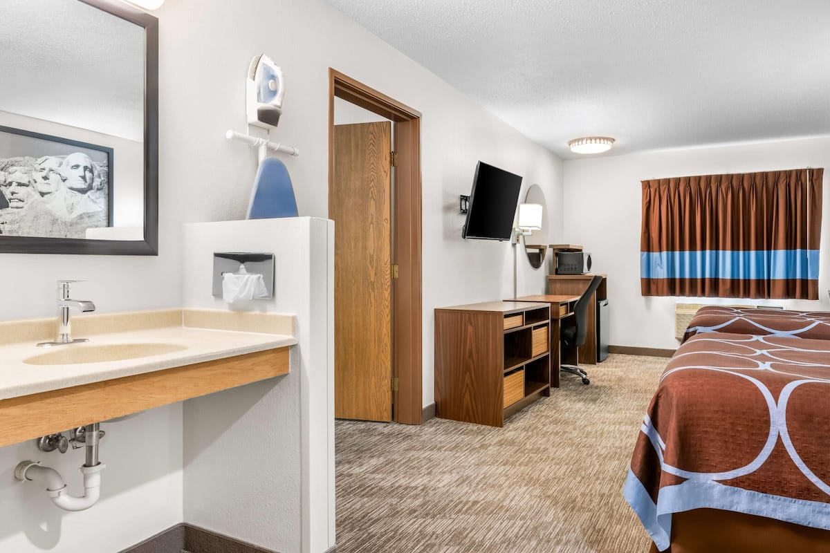 Hotelkamer met een houten bureau, een computer, een televisie, een bed met een decoratieve spreidekking, een badkamer met wastafel en spiegel, en een ingelijst kunstwerk aan de muur.