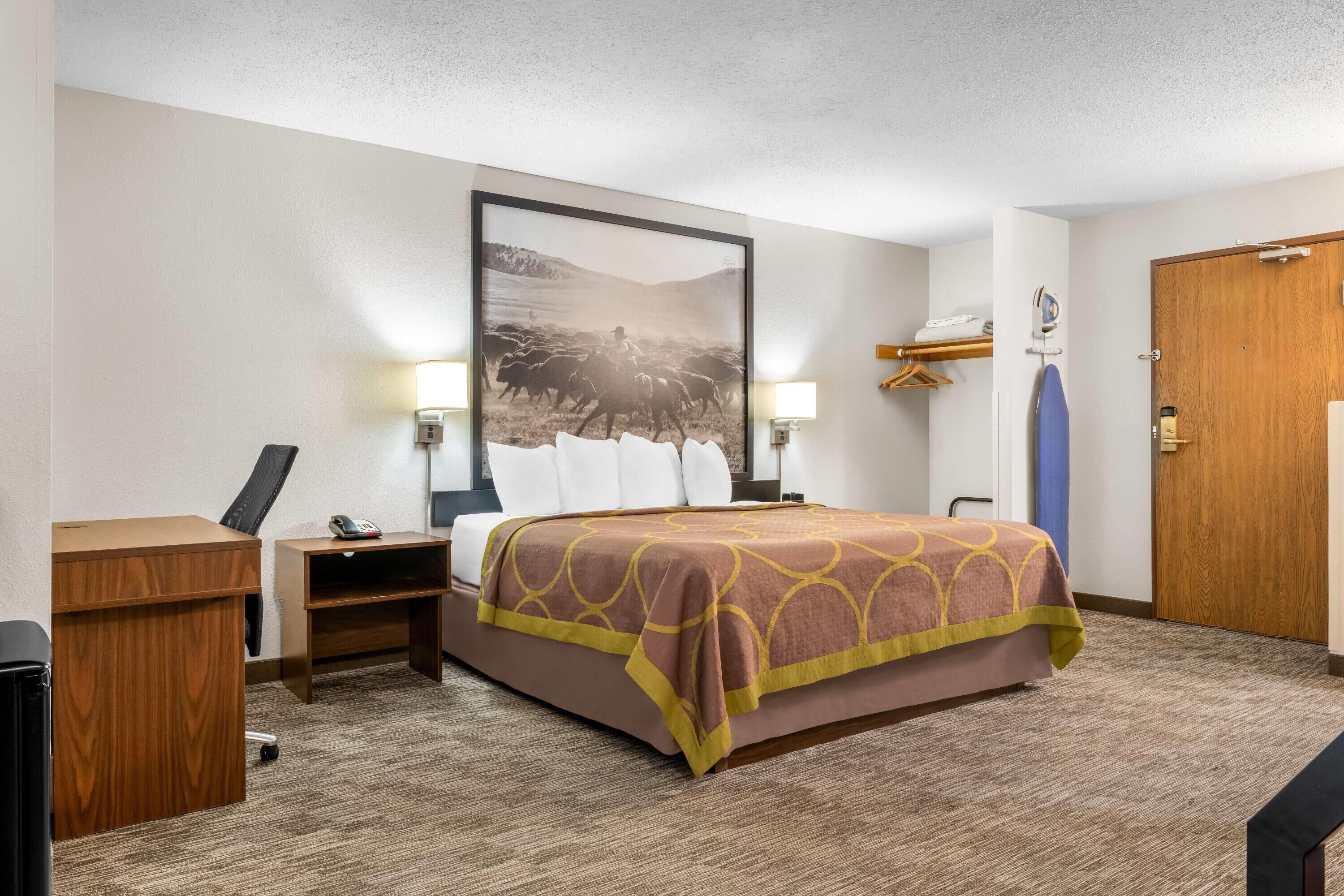 A hotel room with a bed, a desk, a chair, a telephone, a lamp, a door, and a wall-mounted shelf.